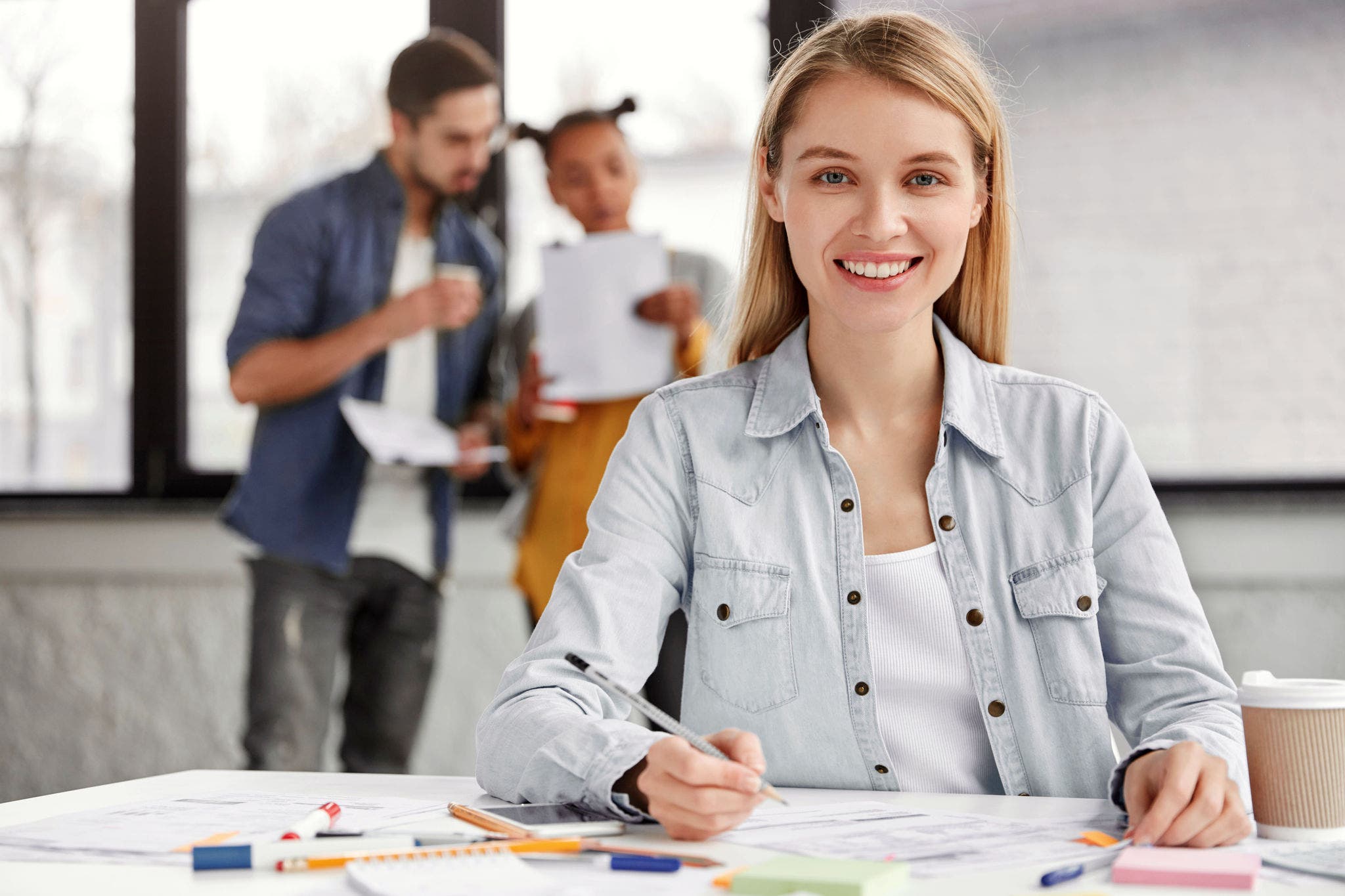 Positive attractive female freelancer work with documentation, sits at coworking space, being in good mood as finishes soon, people discuss something stand in background. Job and occupation concept; Shutterstock ID 1006653625; purchase_order: Dtäubli; job: ; client: ; other: 